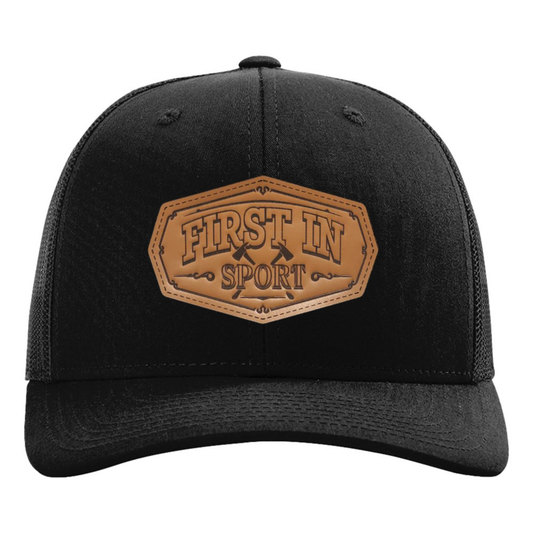 A black trucker-style baseball cap with a brown faux-leather patch on the front that reads 'FIRST IN SPORT'. The cap features black mesh side panels and a curved brim.
